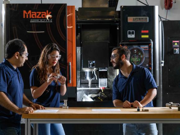 Three individuals standing at a workbench in front of a large Mazak hybrid manufacturing machine, discussing components and technical drawings.