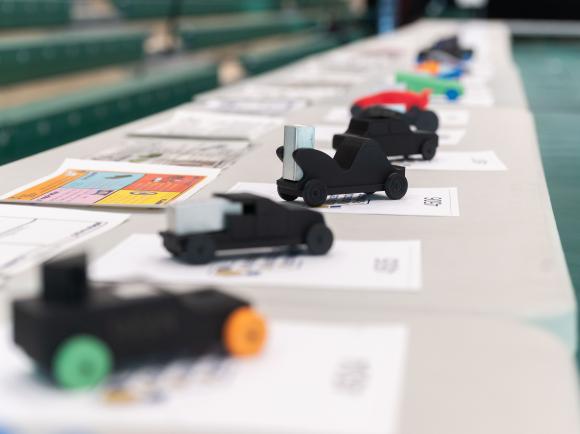 A row of small, student‑designed model race cars displayed on a table, each placed on top of design sheets in a gym setting.